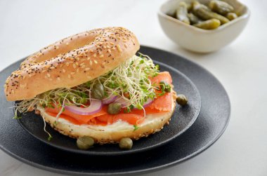 Fresh and healthy smoked salmon on sesame seed bagel with cream cheese, red onion, sprouts and capers on black textured plate in horizontal format.