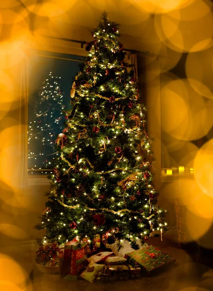 暗闇の中で夜に大きな窓のある家の中で光とプレゼントを持つクリスマスツリーは 焦点を当てて光るライトの外 — ストック写真