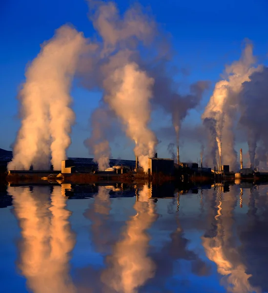 Factory chimneys spewing pollution into the sky smoke rising pollutants in the air reflected in water lake or pond