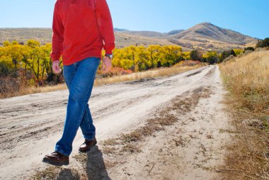Terkedilmiş toprak bir yolda yürüyen ve arka planda dağlar olan ve renkli ağaçlar düşen bir adam.