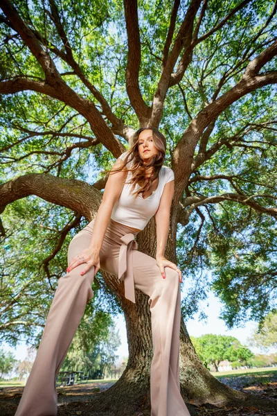 Photo of an attractive young woman crouching down towards camera wiht oak tree looming in background