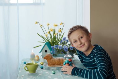 Paskalya pastası, yumurta ve paskalya dekoru olan bir çocuk masada oturuyor.