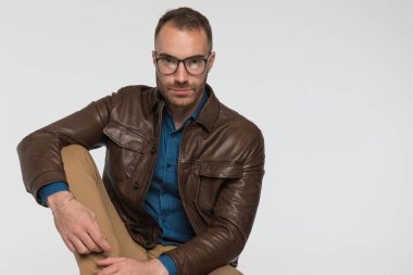 Çekici, sıradan bir adam, bacağını tutarken kameraya bakıyor ve gri stüdyo arka planında kahverengi deri ceket ve gözlük takıyor.