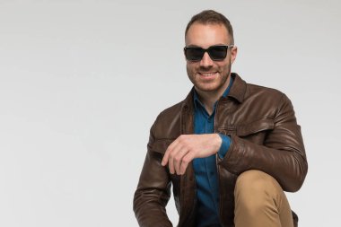 Mutlu sıradan bir adam dirseğini dizinde dinlendiriyor ve gri stüdyo arka planında kahverengi deri ceket ve güneş gözlüğü takıyor.