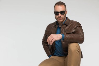Seksi sıradan bir adam patron gibi çömeliyor ve gri stüdyo arka planında kahverengi deri ceket ve güneş gözlüğü takıyor.