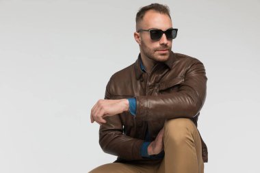 Yakışıklı sıradan bir adam gözlerini kaçırıp gri stüdyo arka planında kahverengi deri ceket ve güneş gözlüğü takıyor.