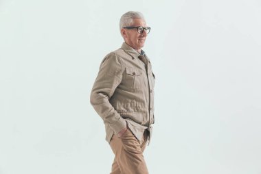 Gri saçlı, elleri ceplerinde yürüyen ve gülümseyen sıradan bir adamın yan görüntüsü.