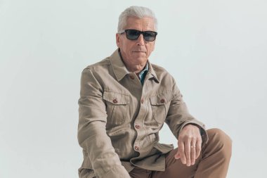 60 'lı yaşlardaki havalı iş adamı güneş gözlüğü takıyor, ellerini kucağında dinlendiriyor ve gri stüdyo arka planında sert bir tavırla poz veriyor.