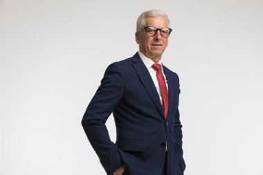 Donanma takım elbiseli yaşlı işadamı ellerini cebine koyup gri stüdyo geçmişine karşı poz veriyor.