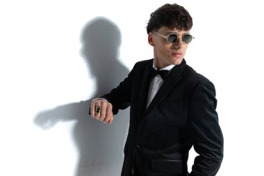 Çekici iş adamı duvara yaslanıyor, başka tarafa bakıyor ve beyaz stüdyo arka planına yüzük ve güneş gözlüğü takıyor.