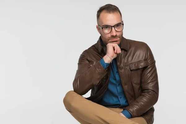 casual man is touching his chin while sitting on a chair and wearing a brown leather jacket and eyeglasses on gray studio background