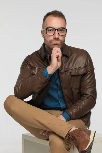 クールなカジュアルな男は脚を交差させながら椅子に座っているグレーのスタジオの背景に茶色の革のジャケットと眼鏡をかけて — ストック写真
