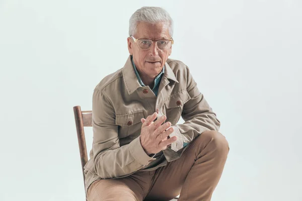 Old businessman rubbing his palms, sitting on a chair and wearing eyeglasses against grey studio background