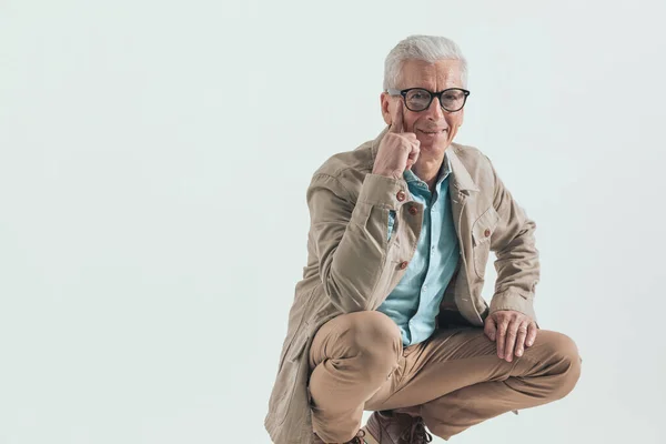 old business man with a smile on his face touching his cheek wearing eyeglasses and squatting on a wooden chair against grey studio background