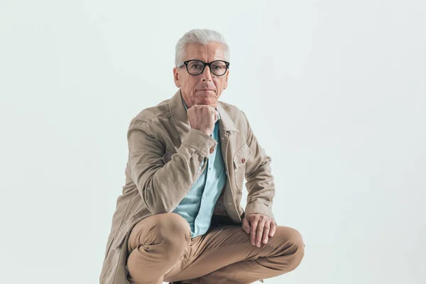 Old businessman leaning his chin in his fist wearing eyeglasses and squatting on a wooden chair against grey studio background