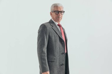 60 'lı yaşlarındaki zarif bir adamın, gri arka planda kırmızı kravatlı, uzun paltolu ve takım elbiseli görüntüsü.
