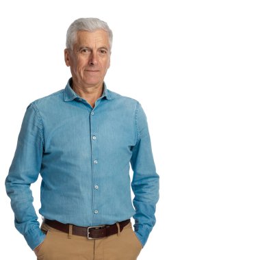 Gri saçlı, genç ruhlu, elleri ceplerinde poz veren ve stüdyoda beyaz arka planda gülümseyen sıradan bir ihtiyar.