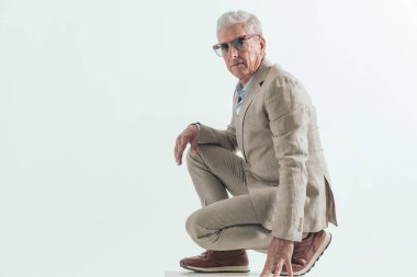 60 'lı yaşlarda güneş gözlüğü takan, çömelmiş, dizlerinin üstüne çömelmiş, stüdyoda gri arka planda poz veren yaşlı bir adamın yan görüntüsü.