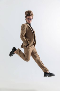 Elleri ceplerinde gezen, havaya atlayan ve stüdyoda gri arka planda poz veren zeki bir adam.