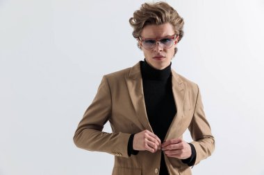 Uzun saçlı, güneş gözlüğü takan, takım elbiseli ve gri arka planda havalı ve ciddi bir şekilde poz veren kendine güvenen moda adamı.