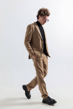 Sarı saçlı, elleri ceplerinde yürüyen ve stüdyoda gri arka planda adımlarına bakan seksi bir genç adam.