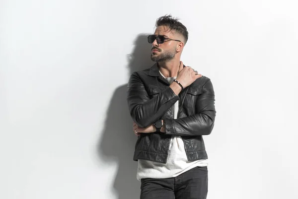 sexy young man in leather jacket with sunglasses looking to side and holding arms in fashion pose in front of grey wall in studio