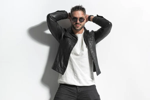 sexy fashion man in leather jacket with sunglasses holding hands behind head and posing in a cool way in front of grey wall in studio
