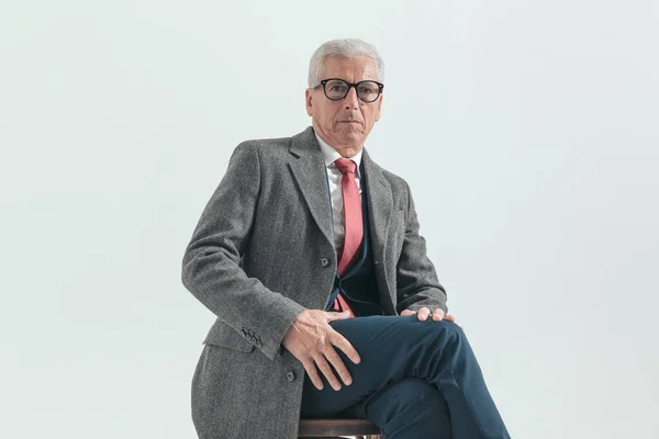 businessman in his 60s with grey hair sitting in a confident way on wooden chair, wearing nice suit and long coat and posing on grey background