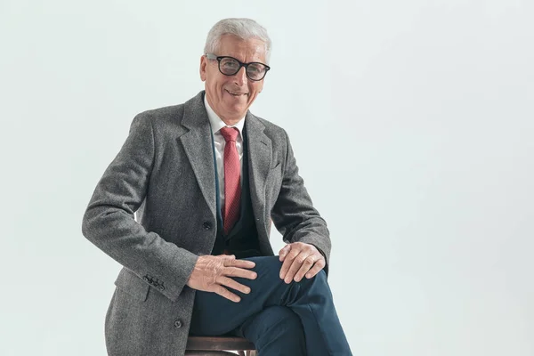 seated man with grey hair wearing suit, long coat and glasses, smiling and being happy on grey background in studio