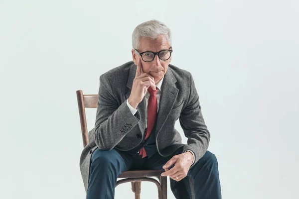 thoughtful man with grizzled hair touching face and thinking, posing with elbows on knees while sitting on grey background in studio