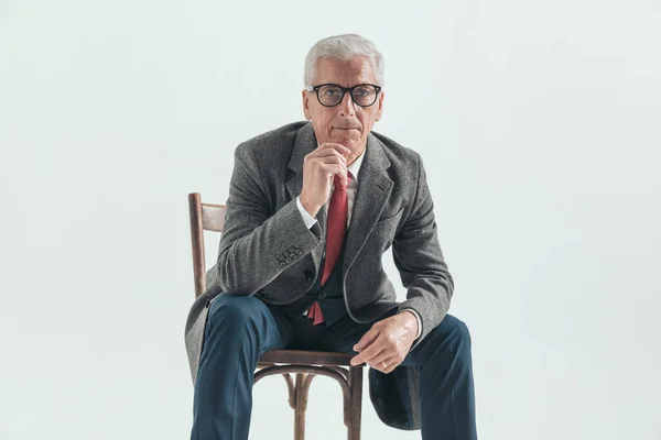 pensive old guy in elegant suit with long coat touching chin and thinking while sitting with elbows on knees on grey background