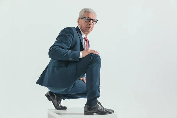 handsome elegant man with grizzled hair in suit crouching and touching knee with palm while posing on grey background