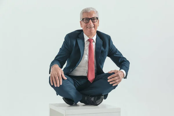 happy old man in his 60s sitting in a turkish pose and laughing while posing in front of grey background