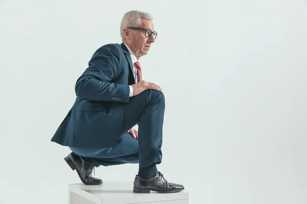 side view of crouched old man in his 60s wearing suit and looking to side while posing in front of grey background in studio