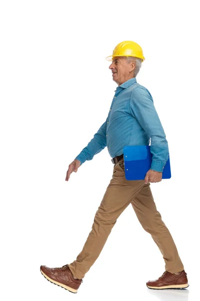 安全黄色のヘルメットとクリップボードの歩行と白の背景の前で笑みを浮かべて誇りに思っている古いエンジニア — ストック写真