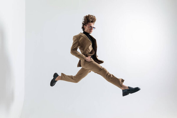 side view of cool fashion man with long hair holding hands in pockets and jumping in the air in front of grey background in studio