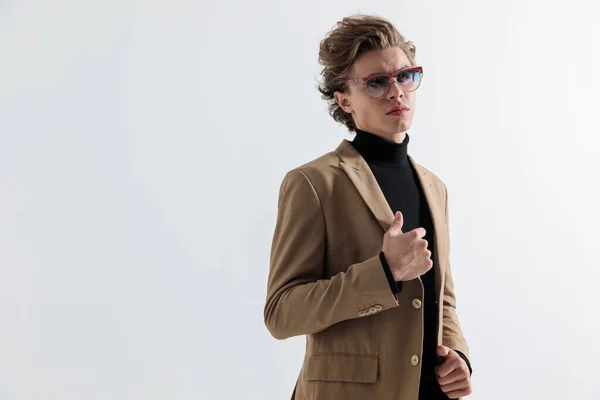 スタジオでグレーの壁の前でスーツを引っ張ったりアレンジしたりしながらサングラスをかけたクールなファッションの男 — ストック写真