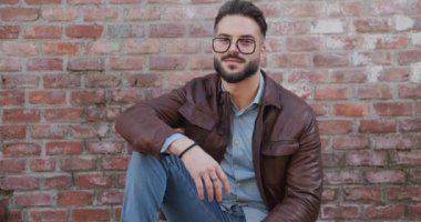 Tuğla duvarın önünde oturan kendine güvensiz adam, dirseğini dizine dayamış, gözlük ve ceketi düzeltiyor, yan tarafa bakıyor ve avuç içini ovuyor.