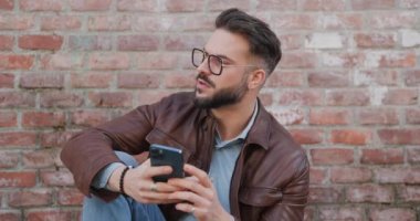Esmer, gözlüklü genç bir adam telefon tutuyor, e-postaları okuyor ve eski bir şehirde dirseğiyle poz verirken gülümsüyor.