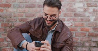 Tıraşsız mutlu bir adam cep telefonunu tutuyor, Instagram 'da kayıyor, gülümsüyor ve ortaçağ şehrinin dışındaki tuğla duvarının önünde otururken yan tarafa bakıyor.