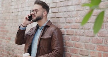 Çekici bir adam kahve içiyor, telefonla konuşurken yan tarafa bakıyor, dışarıda tuğlaların önünde gülümsüyor ve poz veriyor.
