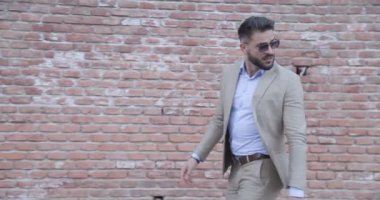 Havalı saç stili olan seksi sakallı adam tuğla duvarın önünde yürüyor, arkasına bakıyor ve koşuyor, dışarıda askeri selamlar yapıyor.