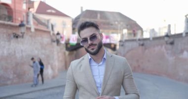 Mutlu genç işadamı güneş gözlüğünü kaldırıyor, gülümsüyor ve eski bir ortaçağ kasabasının önünde gülüyor.