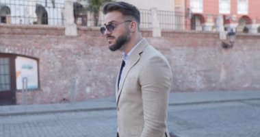 Arkadan bakan, güneş gözlüğü takan, avuçlarını ovuşturan ve eski bir şehirde yürüyüp giden şık bir adamın yan görüntüsü.