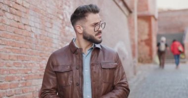 Genç seksi moda adamı etrafa bakıyor, kahverengi deri ceketini ayarlıyor ve Romanya 'nın ortaçağ şehrinin dışında yürürken avuçlarını ovuyor.