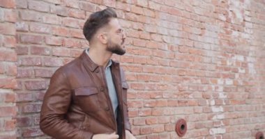 Seksi genç adam duvarda uzanıyor ceketi ve gözlükleri ayarlıyor, yana bakıyor, avuçlarını ovuyor ve gülümsüyor.