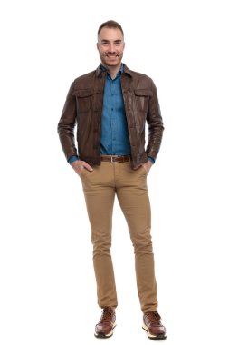 full body picture of handsome man with brown leather jacket smiling and posing with hands in pockets on white background in studio