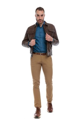 confident young man adjusting brown leather jacket and walking in front of white background in studio