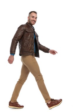 side view of happy guy in brown leather jacket smiling and walking in front of white background in studio