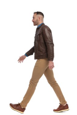 side view of casual bearded man in brown leather jacket walking in front of white background in studio